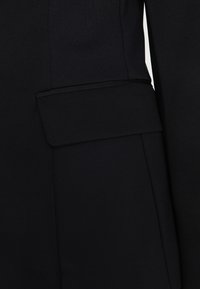 Musta räätälöity takki, jossa on tekstuuripinta, sileä kaulus ja korostettu taskuyksityiskohta hihassa.