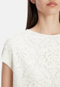 Weißes besticktes Oberteil mit aufwendigen floralen Mustern und strukturierten Verzierungen, runder Ausschnitt und kurzen Ärmeln.