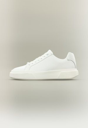 Zapatilla deportiva de perfil bajo totalmente blanca con cierre de cordones, suela gruesa y talón acolchado, mostrada sobre un fondo neutro claro.