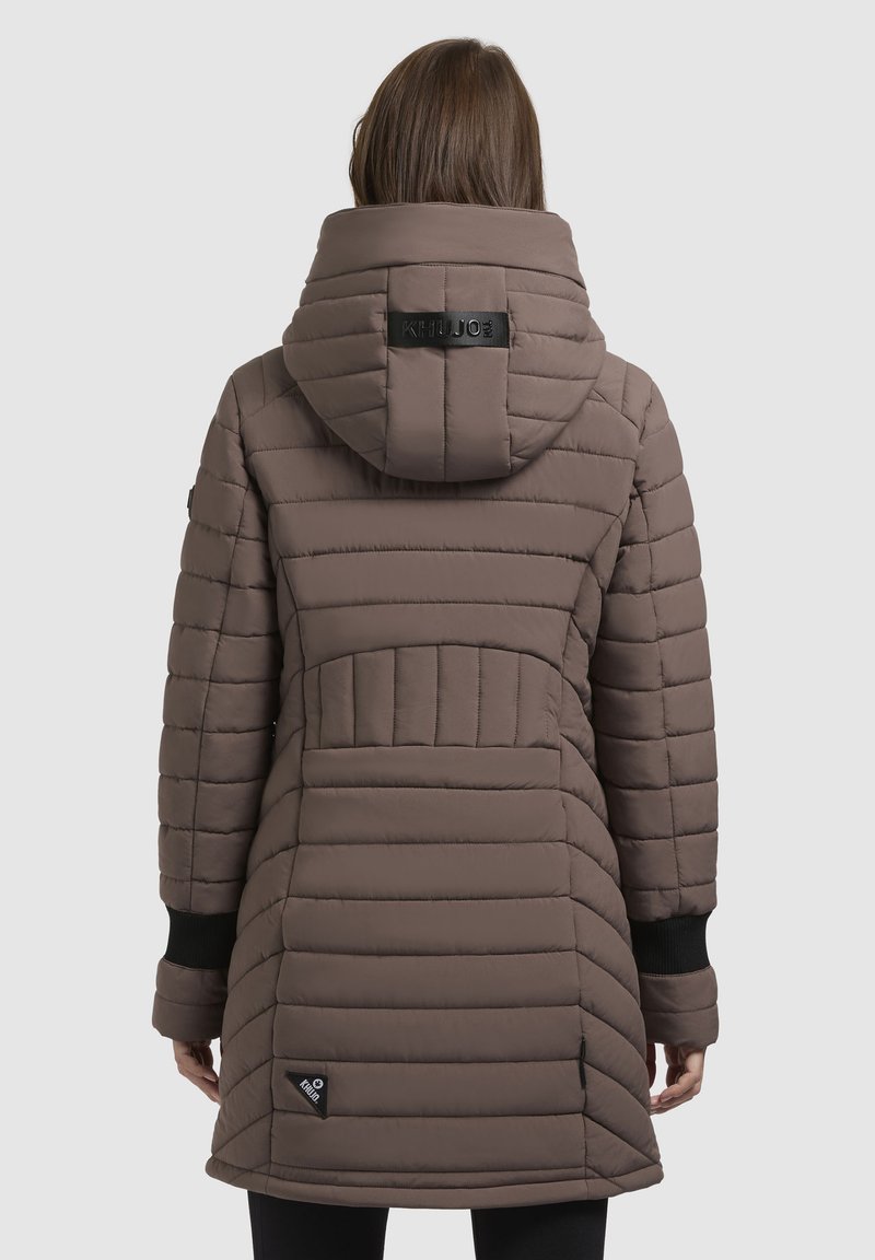 Mujer vestida con un abrigo de invierno largo acolchado en color topo con capucha, puños negros y logotipos de la marca en la capucha y la parte baja de la espalda, de pie de espaldas.