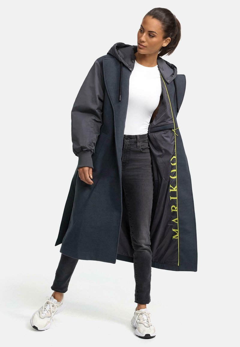 Dark grey long coat with a hood, featuring a contrasting darker fabric and neon yellow lettering inside. Paired with black jeans and white sneakers.