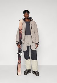 Conjunto de esqui com uma jaqueta puffer cinza claro, camada base cinza escuro e calças de esqui bicolor em cinza e azul-marinho. Botas de esqui pretas e skis incluídos.