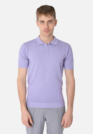 Giovane uomo con capelli corti che indossa una polo a maniche corte viola chiaro e pantaloni grigi, in piedi davanti a uno sfondo bianco semplice.