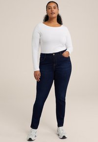 Dunkelblaue hoch taillierte Jeans mit einem Fünf-Taschen-Design, ausgestattet mit goldenen Nähten und einem Knopfverschluss. Kombiniert mit einem weißen Langarm-Top.