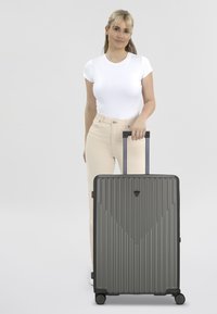 Valise rigide grise avec un design nervuré, ornée d'un logo triangulaire, de poignées noires et de quatre roues pour la mobilité.