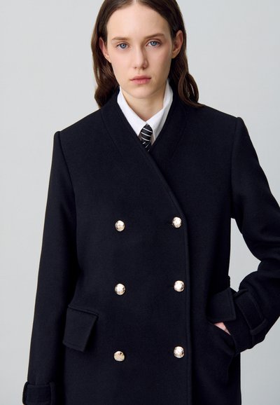Cappotto doppiopetto in lana nera con grandi bottoni argentati, tasche laterali e colletto strutturato. Tessuto liscio e vestibilità sartoriale.