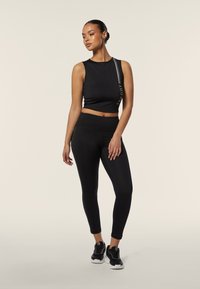 Top corto nero con dettaglio del logo e leggings neri aderenti. Entrambi sono realizzati in un materiale liscio ed elastico. Modello che indossa scarpe da ginnastica nere.
