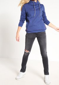 Sudadera azul con capucha y cordones, puños acanalados y bolsillos frontales, combinada con unos vaqueros negros desgastados y zapatillas blancas.