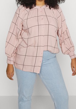 Frau trägt eine lockere rosa Bluse mit schwarzen Gitterlinien und hellblauen Jeans, steht vor weißem Hintergrund.