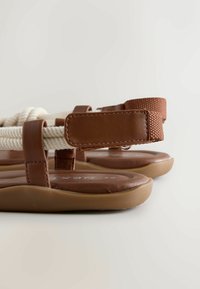 Sandales marron avec semelles matelassées, brides ajustables à scratch et détails en corde blanche sur fond beige.