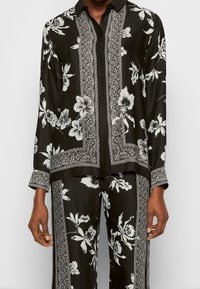 Zwarte bloemenblouse met witte patronen, met een knoopsluiting aan de voorkant, een kraag en contrasterende paisley-bu trim langs de randen en mouwen.