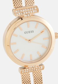 Okrúhly zlatý hodinový strojček so bielym ciferníkom, zlatými hodinovými značkami a sieťovým pásikom. Obsahuje kryštálovú ozdobu na ušiach. Značka: Guess.