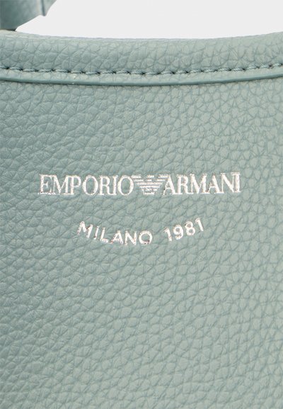 Κοντινό πλάνο υφής ανοιχτόγκρι δέρματος με ασημένιο λογότυπο Emporio Armani και την ένδειξη "Milano 1981" σφραγισμένη από κάτω.