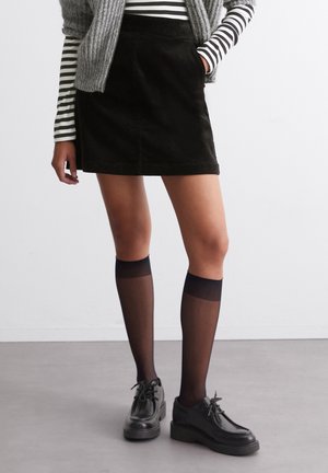Frau trägt einen schwarzen Cord-Minirock, schwarze transparente knielange Strümpfe, schwarze Schnürschuhe und ein gestreiftes Langarmhemd mit grauer Weste.