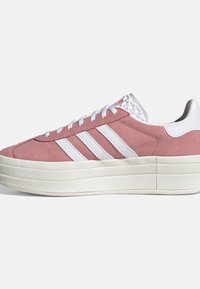 Zapatilla de ante rosa con rayas blancas de Adidas, cordones blancos y suela gruesa y texturizada en un tono blanco roto, sobre un fondo blanco liso.