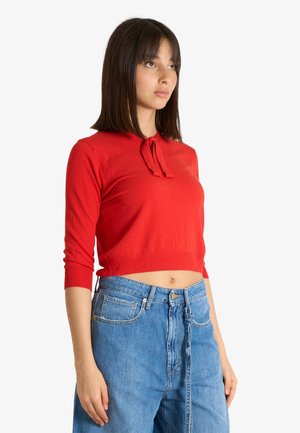 Donna che indossa un top rosso corto a maniche lunghe con dettaglio annodato al collo e jeans blu larghi a vita alta su sfondo bianco.