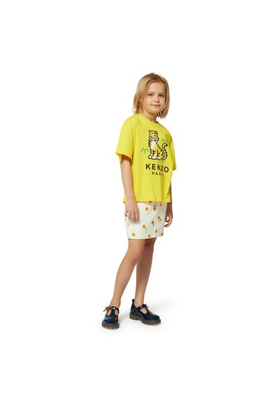 Geel T-shirt met korte mouwen met tijgerprint, gecombineerd met een witte rok met zonnebloempatronen. Draagt donkere schoenen met rode accenten.