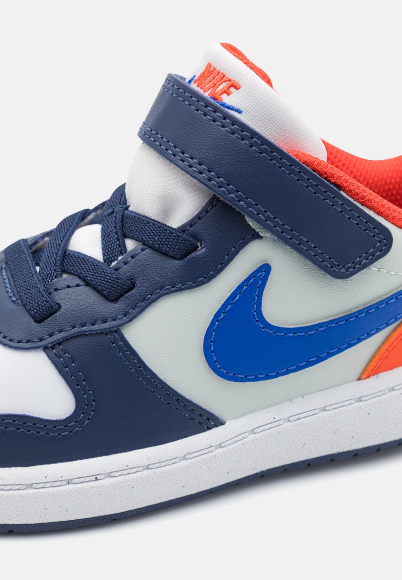 Blau-weiße Sneaker mit einem Klettverschluss, Mesh-Futter und orangefarbenen Akzenten. Strukturierten Gummisohle mit blauem Nike-Swoosh-Logo.
