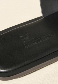 Juodos odinės šlepetės su tekstūruota pado danga, plokščiu padu ir iškaltu logotipu. Lygus paviršius ir minimalistiškas dizainas.
