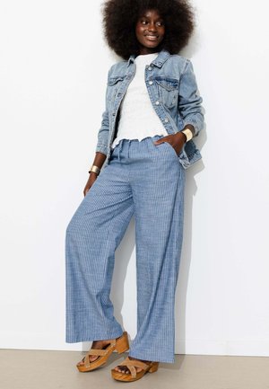 Femme aux cheveux afro portant une veste en denim, un haut blanc texturé, un pantalon large rayé bleu et des sandales compensées en bois, debout et souriante.