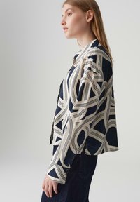 Camicia a righe blu navy e crema con design geometrici, con chiusura a bottoni e maniche lunghe, indossata da un modello di profilo.