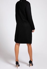 Zwarte jas met een gestructureerd ontwerp, lange mouwen en een knielange snit. Gladde textuur en minimale details. Gekocht met zwarte hoge hakken.