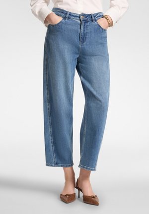Donna che indossa jeans blu a vita alta e a gamba dritta con le mani nelle tasche, abbinati a tacchi marroni a punta e una camicia bianca a maniche lunghe.