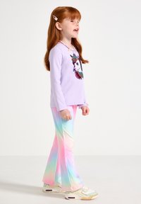 Fille rousse portant un t-shirt licorne, un pantalon flare pastel arc-en-ciel et des baskets, debout face à droite, bouche ouverte comme si elle parlait ou chantait.