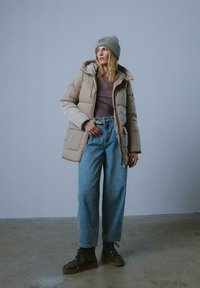 Kurtka puchowa w jasnym beżu, w zestawieniu z purpurową bluzką oraz dżinsami w kolorze niebieskim o wysokim stanie. Beżowa dziana czapka i ciemnozielone buty z wiązaniem.