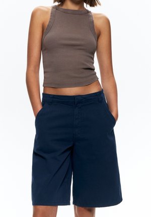 Kvinde iført brun ærmeløs crop top og marineblå knælange shorts med hænderne i lommerne mod hvid baggrund.