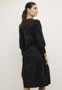 Črna obleka sproščenega kroja, z nabranimi rokavi, okroglim ovratnikom in dolžino do kolen. Narejena je iz gladkega blaga z rahlo teksturo.