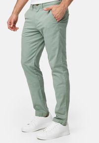 Chinos de algodón verde claro con un ajuste slim, que cuentan con bolsillos laterales y cierre frontal de botón, combinados con zapatillas blancas.