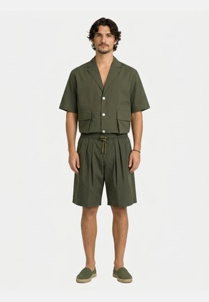 Uomo che indossa una camicia verde oliva a maniche corte con bottoni e tasche frontali, pantaloncini abbinati con coulisse e scarpe slip-on, in piedi su sfondo bianco.