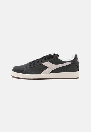 Sort lavtopet sneaker med hvidt sidelogo, sorte snørebånd, hvid sål og hvid polstret krave, vist fra ydersiden på en grå baggrund.