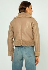 Giacca in pelle marrone tagliata con dettagli in shearling beige sul colletto e sull'orlo. Presenta maniche lunghe e una texture liscia.