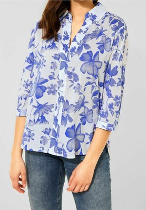 Kvinde iført hvid bluse med blå blomsterprint og 3/4 ærmer, kombineret med blå denimjeans, foran en ensfarvet grå baggrund.