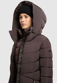 Brown puffer jacket with a large hood, quilted texture, and front zipper. Worn with a black knitted beanie.