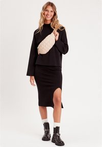 Pull oversized noir, jupe midi noire avec fente sur le côté, sac banane matelassé beige, bottes noires chunky avec détail en chaîne, chaussettes blanches.