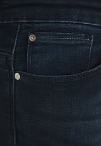 Jeans de denim azul escuro com um design clássico, apresentando um pequeno bolso frontal, acessórios em prata e detalhes de costura discretos.