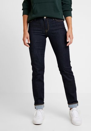 Personne portant un jean bleu foncé retroussé, des baskets blanches et un sweat à capuche vert foncé, debout devant un fond uni.
