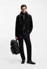 Chaqueta negra con cremallera y cuello de piel sintética, jersey de cuello alto gris, pantalones oscuros y mocasines negros. Sosteniendo una mochila de cuero verde.