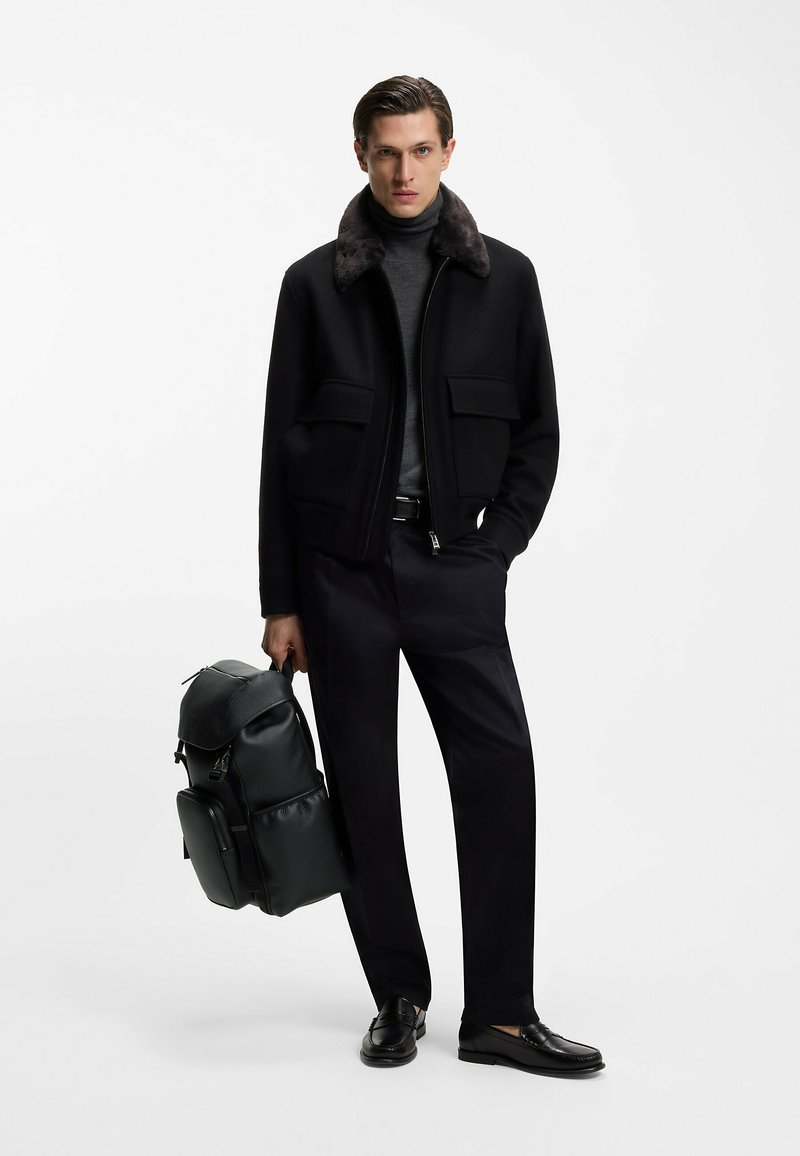 Chaqueta negra con cremallera y cuello de piel sintética, jersey de cuello alto gris, pantalones oscuros y mocasines negros. Sosteniendo una mochila de cuero verde.