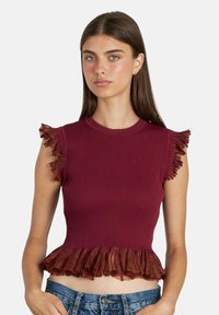 Giovane donna con capelli castani lisci che indossa un top senza maniche a coste bordeaux con rifiniture arricciate e jeans blu, sfondo neutro.