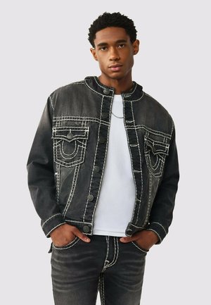 Jeune homme portant une veste en denim noire avec des surpiqûres blanches sur une chemise blanche, les mains dans les poches, debout devant un fond gris clair uni.