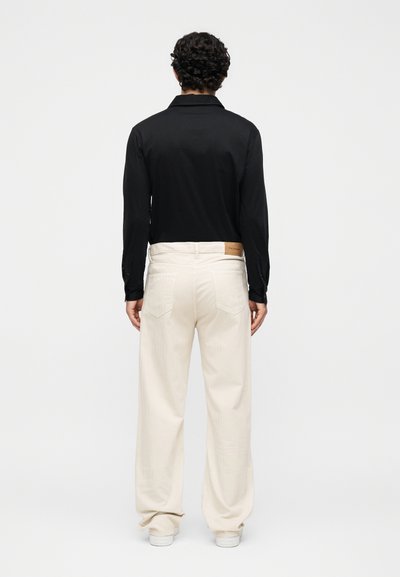 Άντρας με κατσαρά μαλλιά που φοράει μαύρο πουκάμισο μακρυμάνικο και φαρδιά λευκή παντελόνα, στέκεται αντικρίζοντας ένα απλό ανοιχτό φόντο.