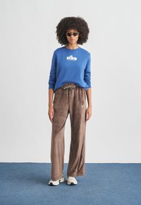 Γυναίκα που φορά μπλε φούτερ Nike, καφέ βελουτέ παντελόνα με φαρδιά μπατζάκια, λευκά αθλητικά παπούτσια και γυαλιά ηλίου cat-eye, στέκεται πάνω σε μπλε χαλί.