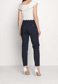 Žena oblečená v námořnických úzkých kalhotách s vroubkovaným pasem, bílé bezrukávové topu a bílých sandálech na vysokém podpatku, stojící na bílém pozadí.