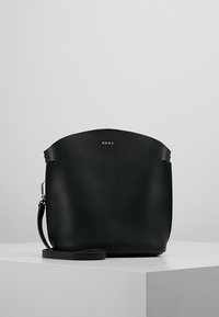 Czarna skórzana torebka crossbody DKNY z wąskim paskiem, złotymi akcentami i zaokrągloną krawędzią u góry, umieszczona na białym podium.