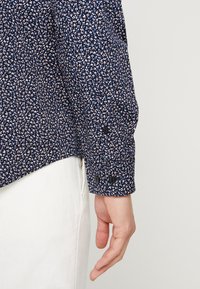 Námořnická modrá košile s malým květinovým vzorem v růžové a světle modré. Má manžety na knoflíky a hladkou strukturu látky.