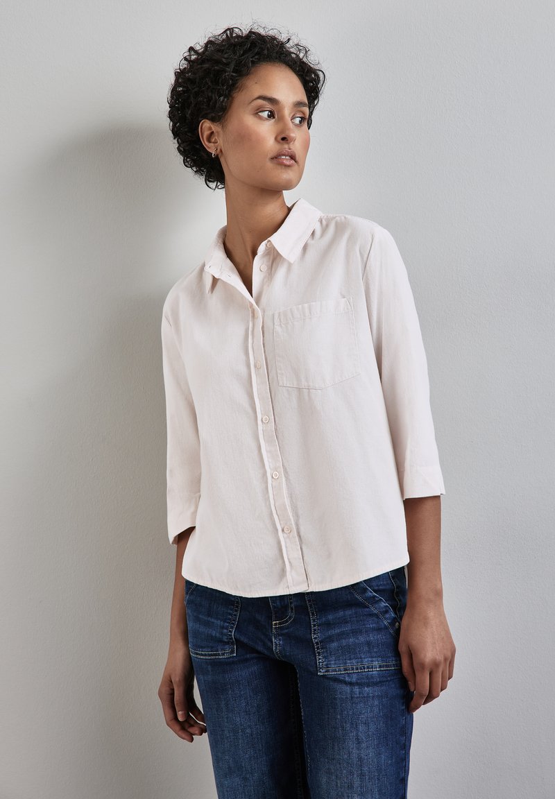 Light pink button-up shirt with a collar, three-quarter sleeves, and a front pocket, paired with dark blue denim jeans.